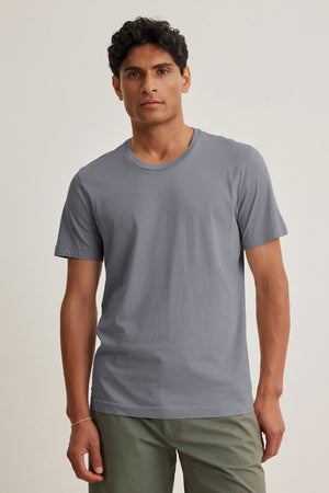 A man faces the camera against a neutral background, wearing the HOWARD TEE by Velvet by Graham & Spencer—a plain grey lightweight Pima jersey t-shirt—paired with green pants.