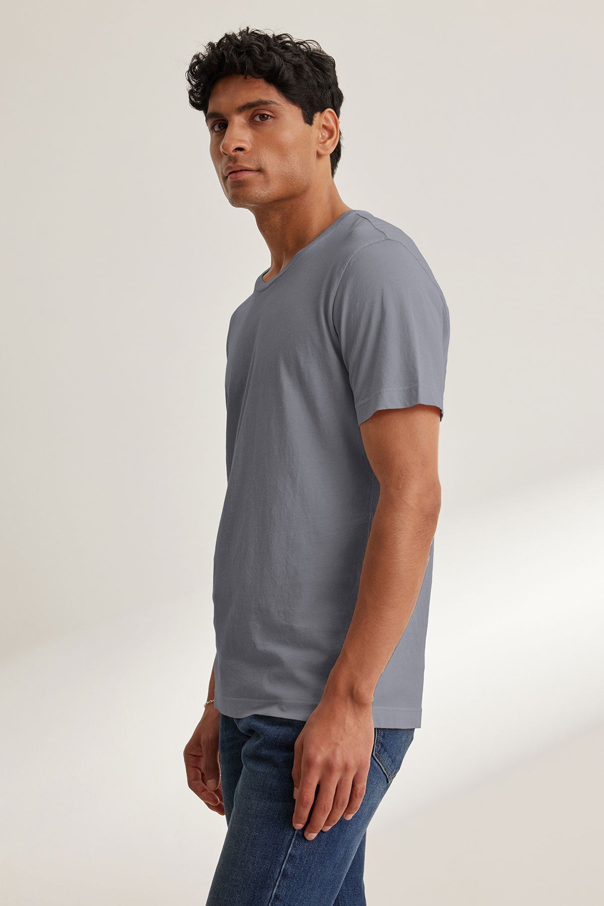 A man stands sideways, wearing the Velvet by Graham & Spencer HOWARD TEE— a plain gray lightweight Pima jersey short-sleeve t-shirt— and blue jeans, looking toward the camera against a neutral background.-40117592359105
