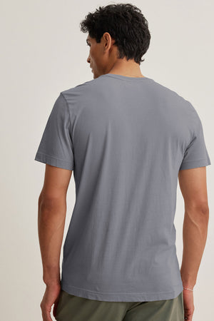 A person stands facing away, wearing the short-sleeved HOWARD TEE in lightweight Pima jersey by Velvet by Graham & Spencer and olive green pants against a plain light background.