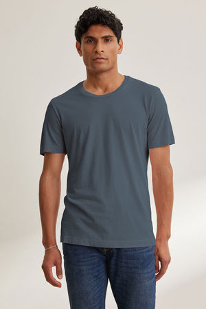 A man faces forward in the Velvet by Graham & Spencer HOWARD TEE, a lightweight Pima jersey short-sleeve grayish-blue T-shirt paired with blue jeans, set against a neutral background for a perfect fit and subtle vintage feel.