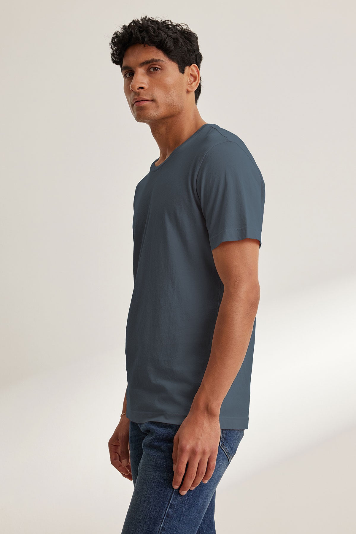 A man stands sideways, wearing the HOWARD TEE by Velvet by Graham & Spencer in dusty blue Pima jersey for a perfect fit, paired with blue jeans against a plain light background.-40117588361409