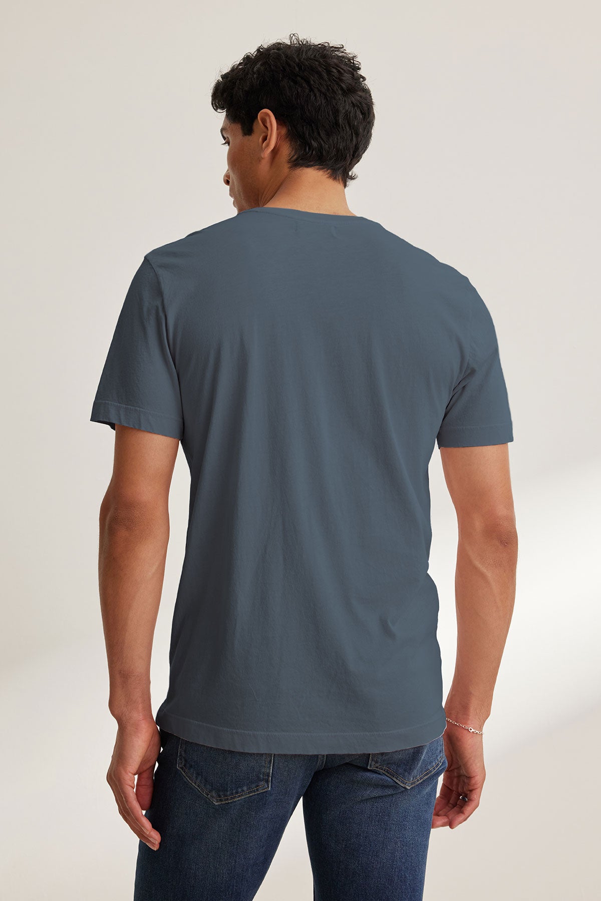 A man faces away from the camera, wearing the HOWARD TEE by Velvet by Graham & Spencer—a plain, lightweight Pima jersey short-sleeve blue t-shirt—paired with blue jeans against a neutral background.-40117588328641