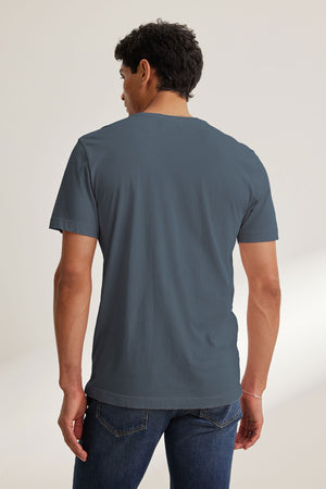 A man faces away from the camera, wearing the HOWARD TEE by Velvet by Graham & Spencer—a plain, lightweight Pima jersey short-sleeve blue t-shirt—paired with blue jeans against a neutral background.