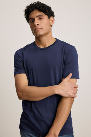 A person with short curly hair stands facing the camera, one arm crossed, wearing Velvet by Graham & Spencer’s HOWARD TEE in plain navy blue lightweight Pima jersey and jeans.