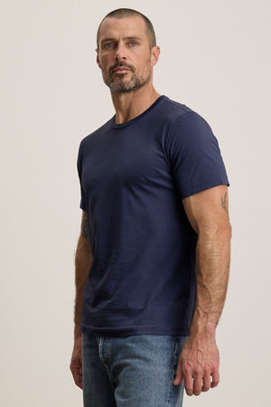 A man with short hair and a beard wears the HOWARD TEE by Velvet by Graham & Spencer—a plain navy lightweight Pima jersey t-shirt—and blue jeans, standing against a neutral background.