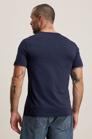 A man with short hair and tattoos stands with his back to the camera, wearing a navy HOWARD TEE by Velvet by Graham & Spencer in lightweight Pima jersey, paired with blue jeans.