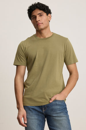A man wears the HOWARD TEE by Velvet by Graham & Spencer, a plain olive green Pima jersey t-shirt, paired with blue jeans. He stands against a neutral background with one hand in his pocket, showcasing its perfect fit.