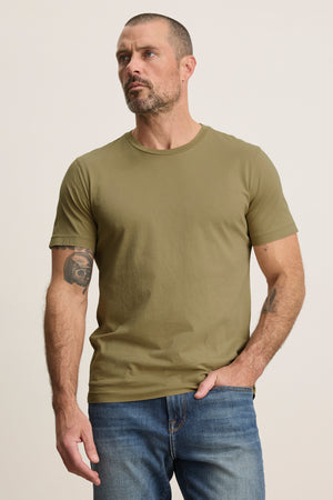 A man with short hair and a beard wears the Velvet by Graham & Spencer HOWARD TEE in olive green and vintage-style blue jeans, standing against a plain light background.