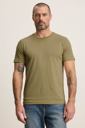 A man with short hair and a beard wears the olive green HOWARD TEE by Velvet by Graham & Spencer with blue jeans, standing against a plain background.