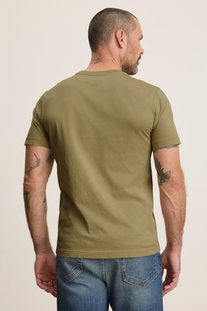 A man with short hair and tattooed arms stands facing away, wearing the Velvet by Graham & Spencer HOWARD TEE in olive green and blue jeans against a plain background for a vintage look.