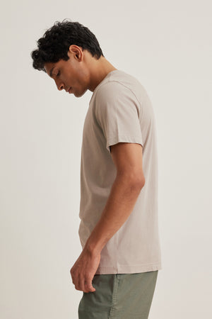 In a simple setting, someone wearing the HOWARD TEE by Velvet by Graham & Spencer in beige Pima jersey and green pants stands facing left, head slightly tilted down, showcasing a perfect fit with a vintage vibe.