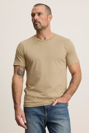 A man with short hair and a beard wears the beige HOWARD TEE by Velvet by Graham & Spencer with blue jeans, standing against a light background. A tattoo on his upper left arm adds vintage flair to this classic look.