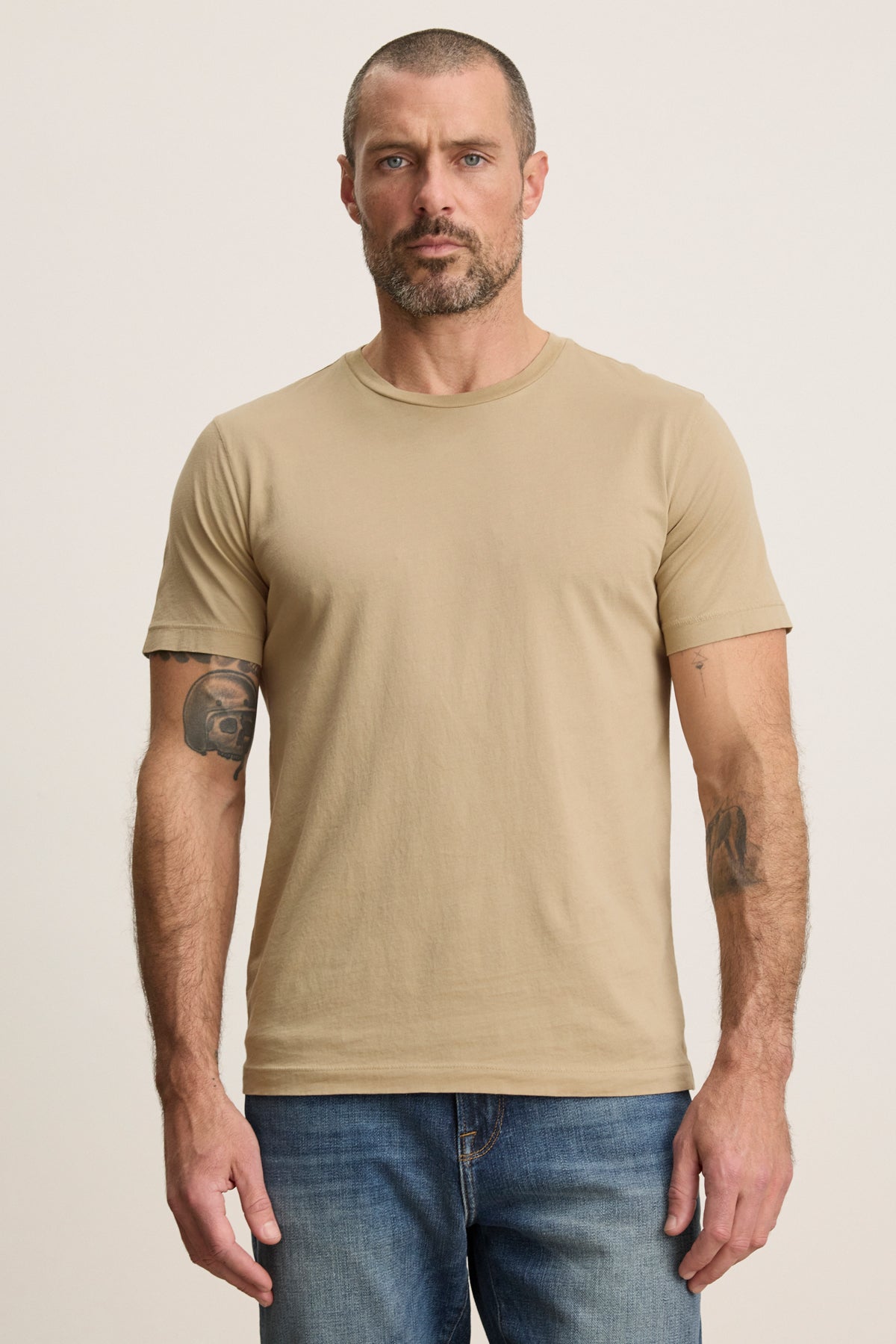 A man with short hair, a beard, and tattooed arms wears the beige HOWARD TEE by Velvet by Graham & Spencer and blue jeans, standing against a plain light background.