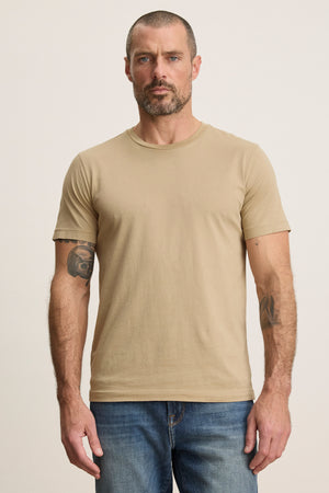 A man with short hair, a beard, and tattooed arms wears the beige HOWARD TEE by Velvet by Graham & Spencer and blue jeans, standing against a plain light background.
