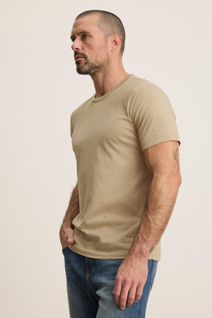 A man with short hair and a beard wears the HOWARD TEE by Velvet by Graham & Spencer in beige, paired with blue jeans, standing against a light background for a perfect fit and vintage feel.