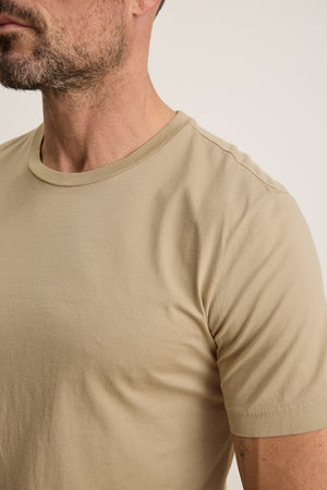 Close-up of a man wearing the HOWARD TEE by Velvet by Graham & Spencer in beige Pima jersey, featuring his neck, partial face, and upper torso against a plain background for a vintage vibe.