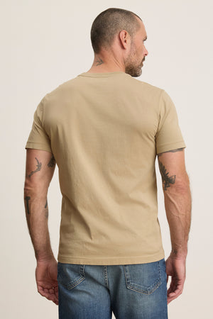 A man with short hair, viewed from behind, wears the beige HOWARD TEE by Velvet by Graham & Spencer and blue jeans. He has several arm tattoos and a small neck tattoo.