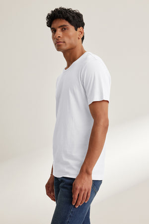 A man stands against a plain background wearing the Velvet by Graham & Spencer HOWARD TEE, made from lightweight Pima jersey, and blue jeans, looking slightly to the side.