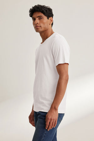 A man stands slightly to the side, wearing the HOWARD TEE by Velvet by Graham & Spencer—a plain white Pima jersey short-sleeve shirt—paired with blue jeans against a neutral background, capturing the perfect fit and vintage feel.