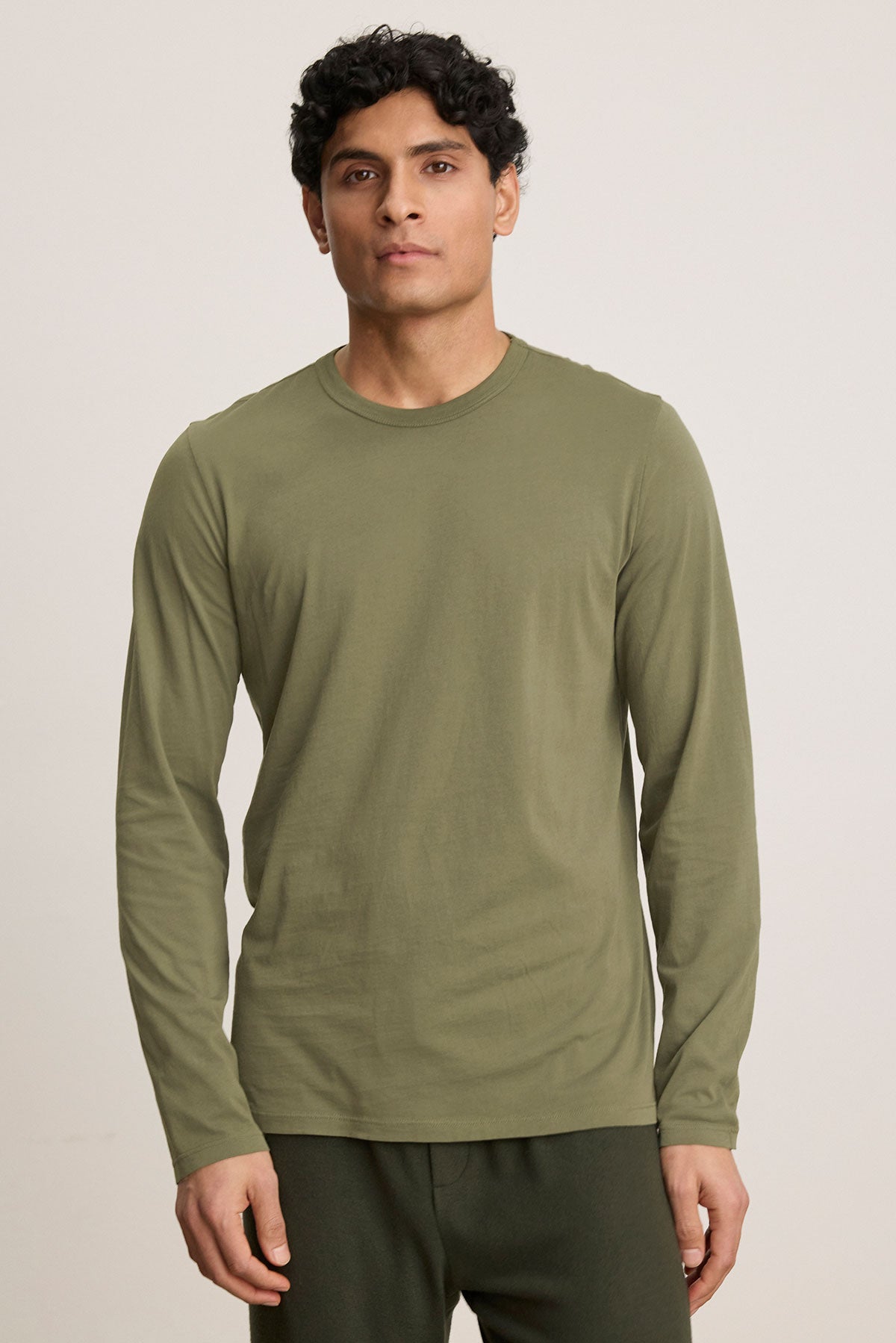 A person wears the Velvet by Graham & Spencer ROWLAND TEE in olive green with dark green pants, standing against a plain light background.