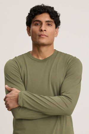 Man with short curly hair stands with arms crossed, wearing the ROWLAND TEE by Velvet by Graham & Spencer—an olive green Pima jersey long-sleeve crewneck—against a plain light background.