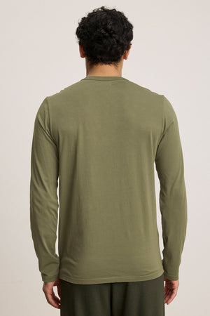 A man with short curly hair is shown from the back, wearing Velvet by Graham & Spencer’s ROWLAND TEE in olive green and dark pants, standing against a plain light background.