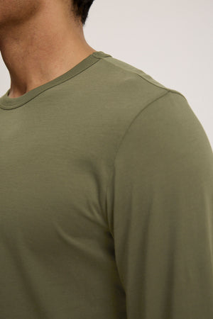 A close-up shows a person wearing the Velvet by Graham & Spencer ROWLAND TEE in olive green Pima jersey, highlighting the left shoulder, crew neckline, and upper chest against a plain background.
