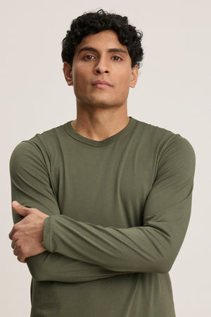 A man with short curly hair stands with arms crossed, wearing the ROWLAND TEE by Velvet by Graham & Spencer—an olive green long-sleeve shirt with a classic crew neckline—looking directly at the camera against a plain background.