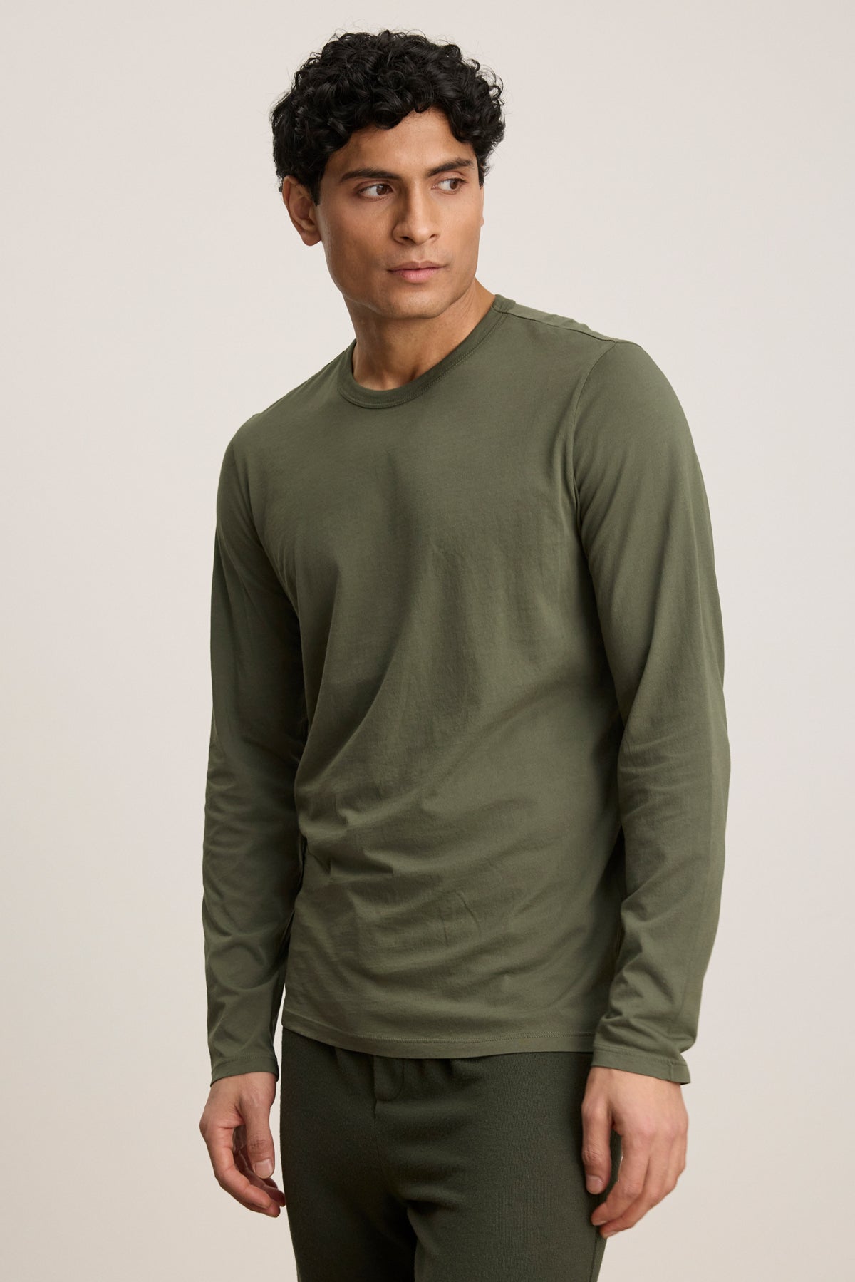 Wearing the ROWLAND TEE by Velvet by Graham & Spencer in olive green with long sleeves and a classic crew neckline, a person pairs it with matching pants and poses against a plain light background, gazing slightly to the side.-40054349267137