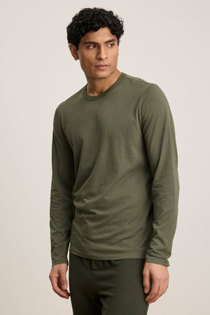 Wearing the ROWLAND TEE by Velvet by Graham & Spencer in olive green with long sleeves and a classic crew neckline, a person pairs it with matching pants and poses against a plain light background, gazing slightly to the side.