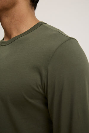 Close-up of a person wearing the ROWLAND TEE by Velvet by Graham & Spencer—an olive green long-sleeve crew neck tee—showcasing the upper chest, shoulder, and neck against a plain background.