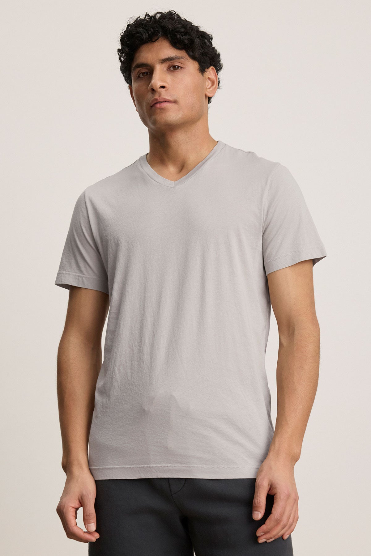 A person stands in a SAMSEN TEE by Velvet by Graham & Spencer, a light gray V-neck crafted from soft Pima jersey. They pair it stylishly with dark pants against a plain background, perfect for layering.