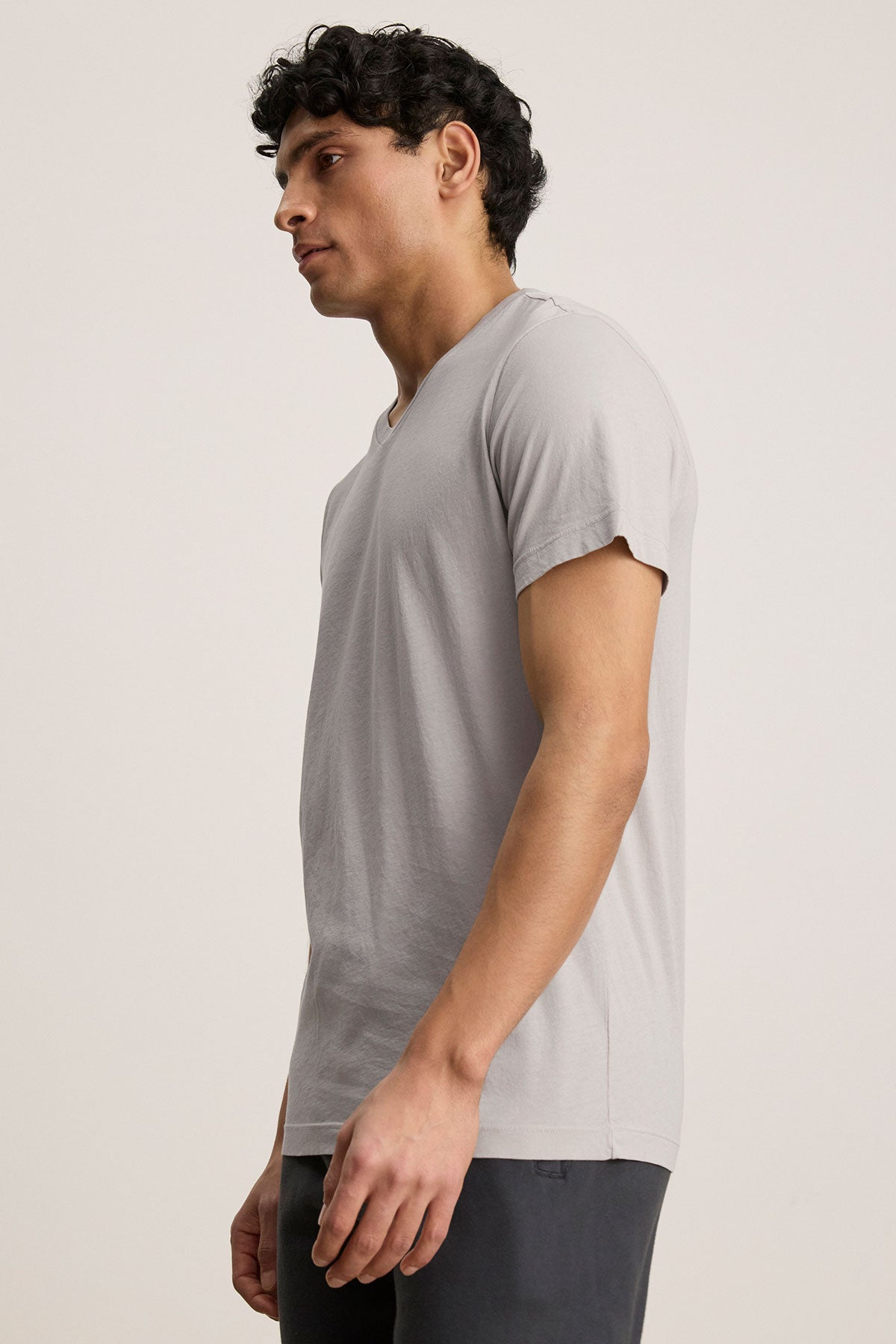 Wearing a Velvet by Graham & Spencer SAMSEN TEE in light gray paired with dark pants, a person stands sideways against a plain backdrop, effortlessly showcasing the art of layering.