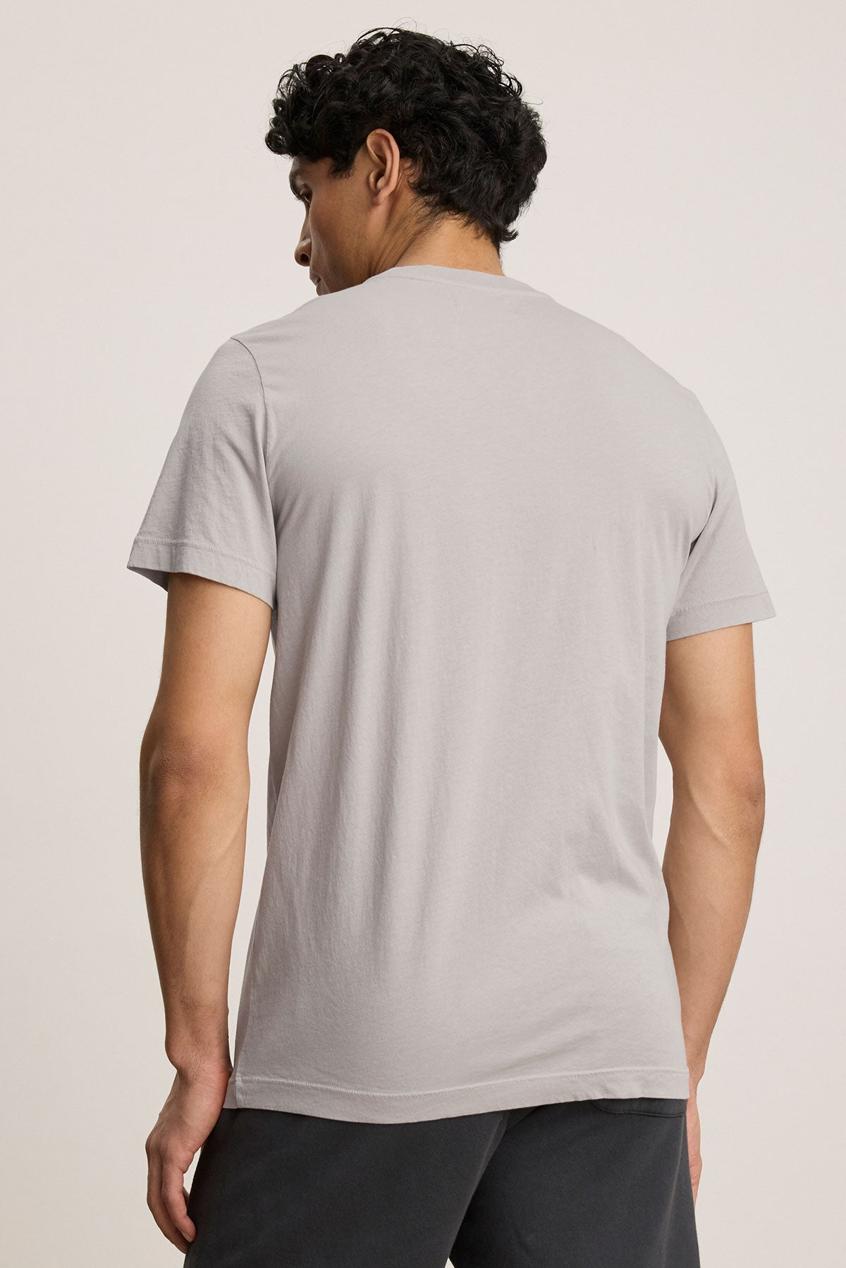A person with short curly hair, wearing a SAMSEN TEE by Velvet by Graham & Spencer in light grey and dark pants, stands with their back to the camera.-38942945804481