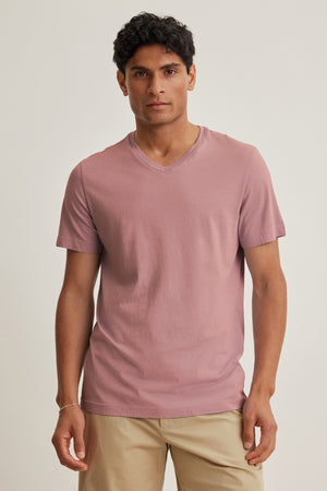 A man faces forward in a Velvet by Graham & Spencer SAMSEN TEE, a short-sleeve mauve Pima jersey V-neck, paired with beige pants against a plain light background.