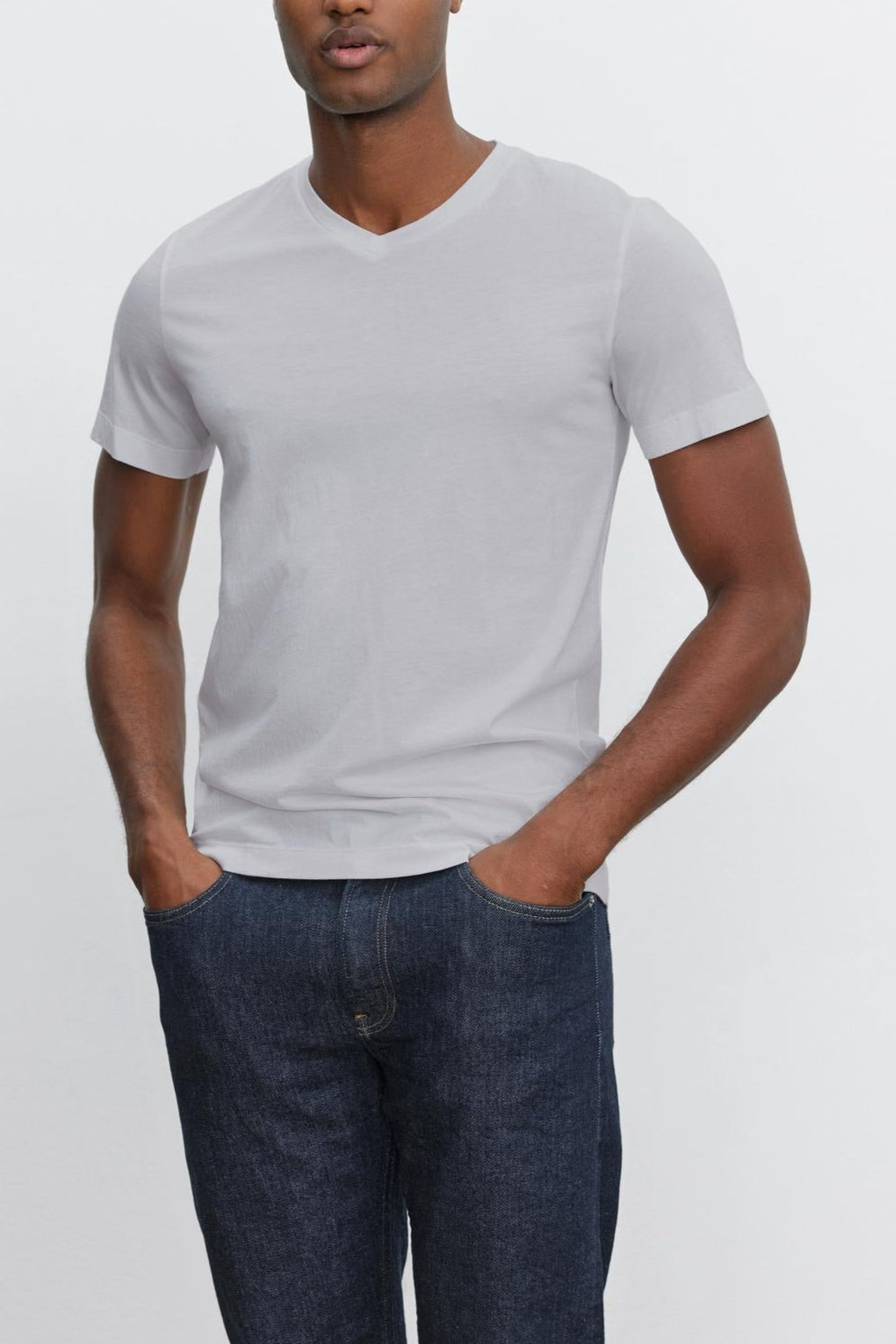 A person is standing with their hands in their pockets, wearing a plain white Velvet by Graham & Spencer SAMSEN TEE with a V-neckline and dark blue jeans—perfect for everyday wear—against a plain white background.