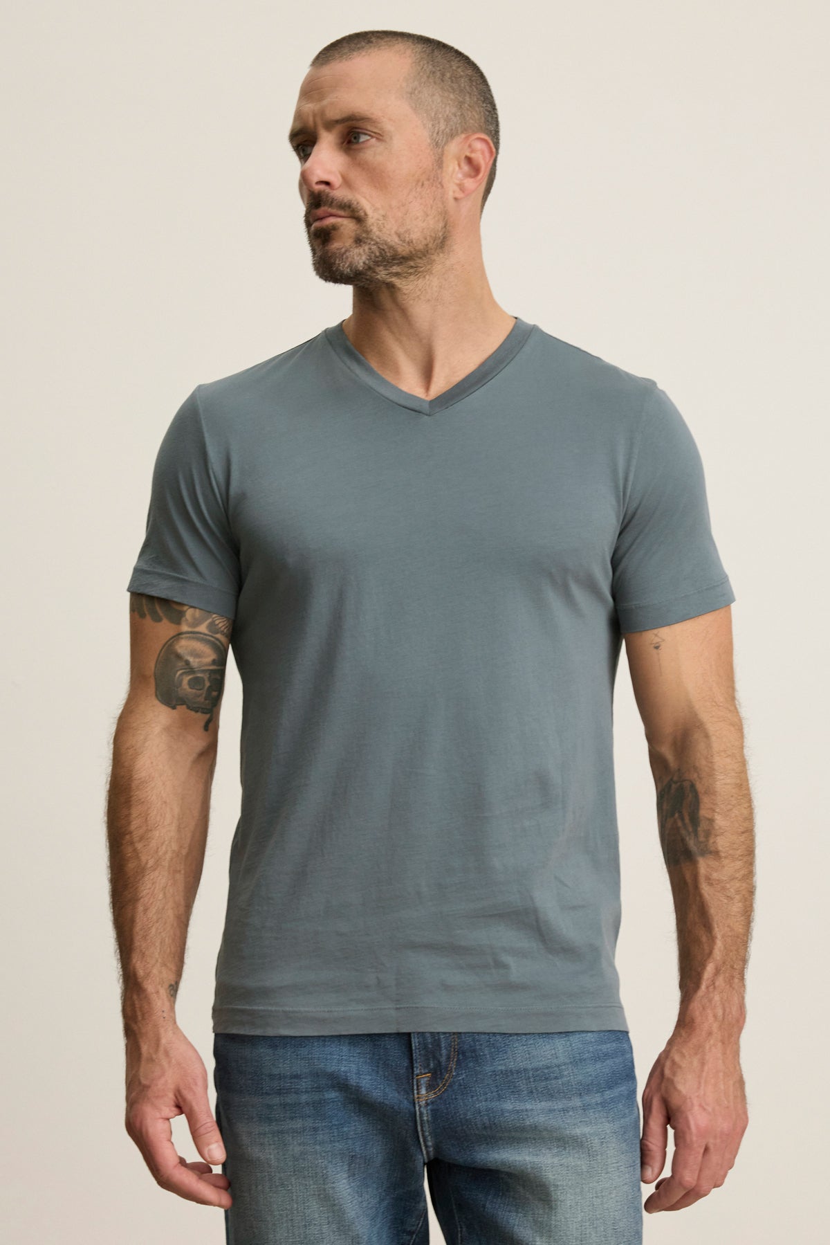 A man with short hair and arm tattoos wears the Velvet by Graham & Spencer SAMSEN TEE, a gray Pima jersey V-neck t-shirt, and blue jeans, standing against a plain light background.-39446046900417