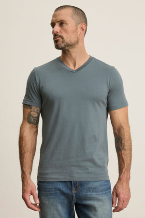 A man with short hair and arm tattoos wears the Velvet by Graham & Spencer SAMSEN TEE, a gray Pima jersey V-neck t-shirt, and blue jeans, standing against a plain light background.
