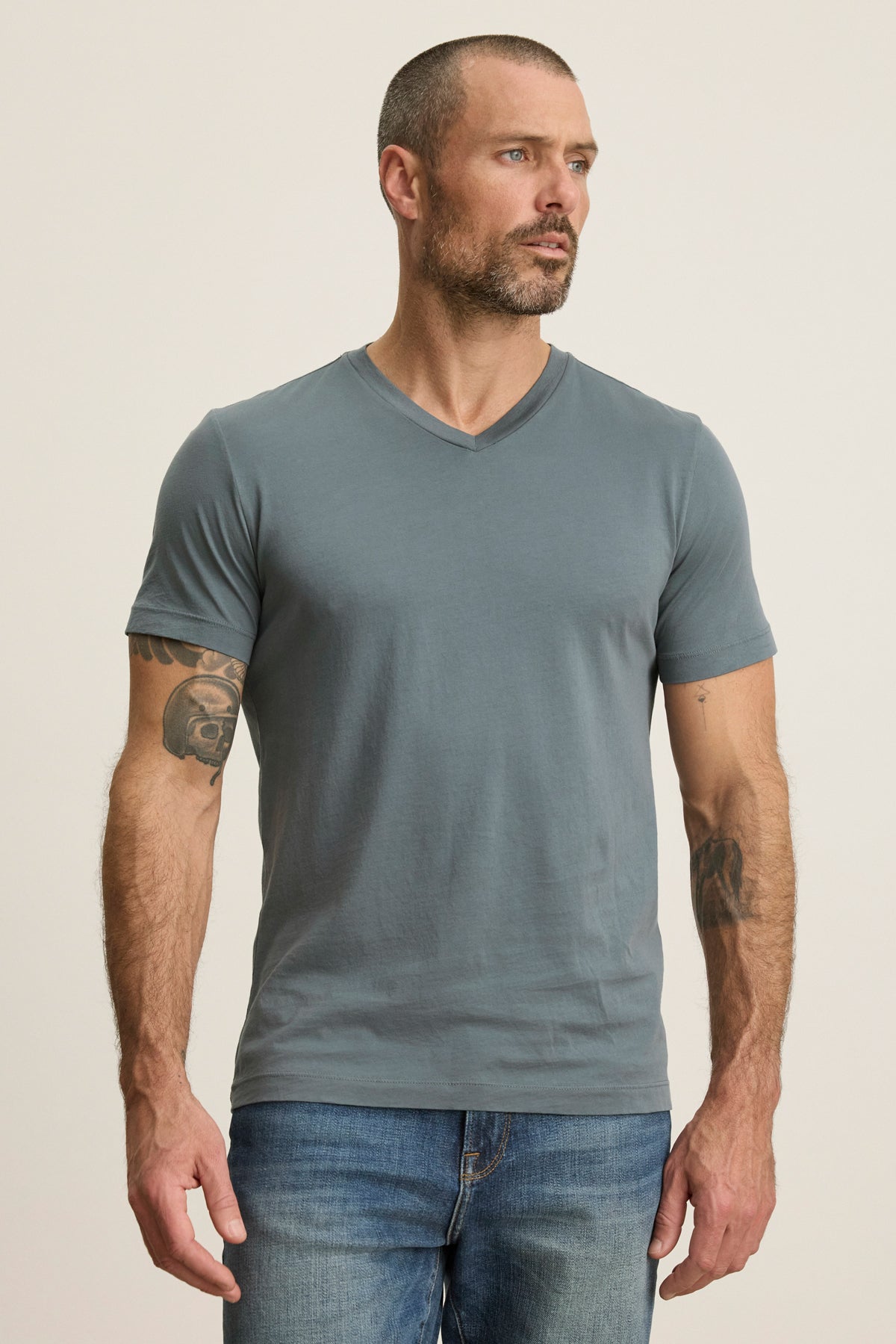 A man with short hair and a beard wears the SAMSEN TEE by Velvet by Graham & Spencer, a grey V-neck Pima jersey T-shirt, paired with blue jeans. He stands against a plain light background with arms relaxed at his sides.-39446046933185