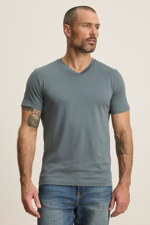 A man with short hair and a beard wears the SAMSEN TEE by Velvet by Graham & Spencer, a grey V-neck Pima jersey T-shirt, paired with blue jeans. He stands against a plain light background with arms relaxed at his sides.