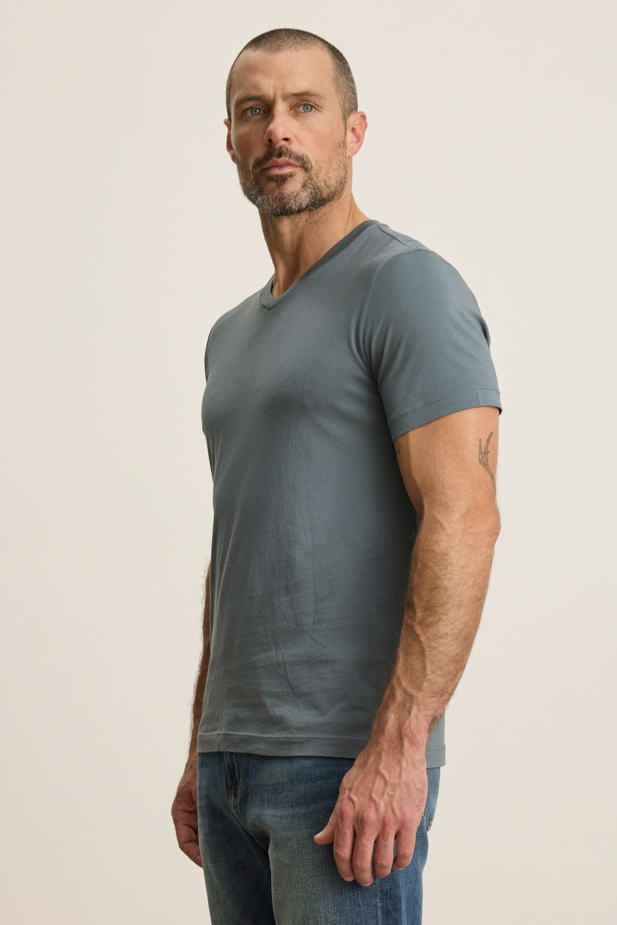 Man with short hair and beard wears the SAMSEN TEE by Velvet by Graham & Spencer—a fitted gray Pima jersey v-neck—paired with blue jeans against a plain light background, facing slightly to the side. Perfect for stylish layering.