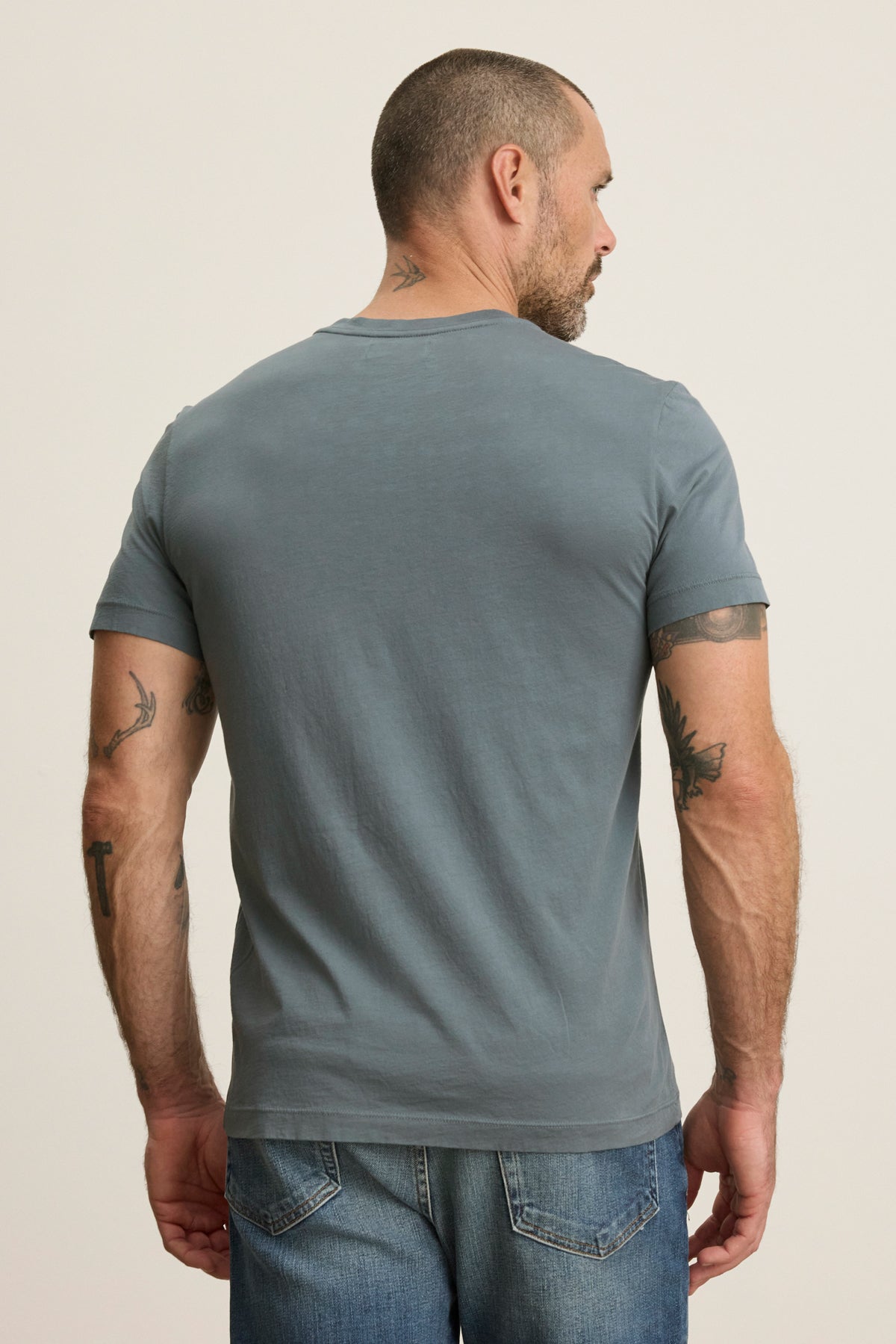 A man with short hair and visible arm and neck tattoos is seen from behind wearing the gray SAMSEN TEE by Velvet by Graham & Spencer and blue jeans.-39446046998721