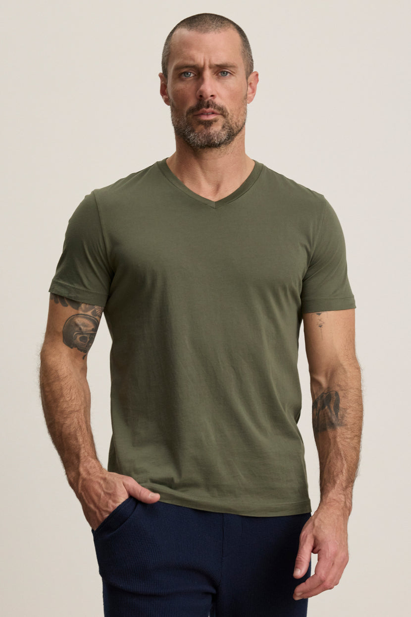 A man with short hair and a beard wears the Velvet by Graham & Spencer SAMSEN TEE in olive green and dark pants, standing before a plain light background—a versatile choice for everyday wear.