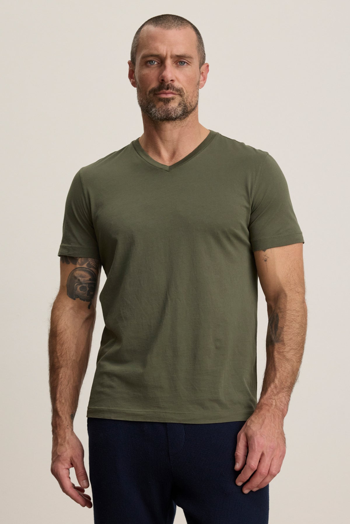 A man with short hair and a beard wears the olive green SAMSEN TEE by Velvet by Graham & Spencer and dark pants, standing against a plain light background.