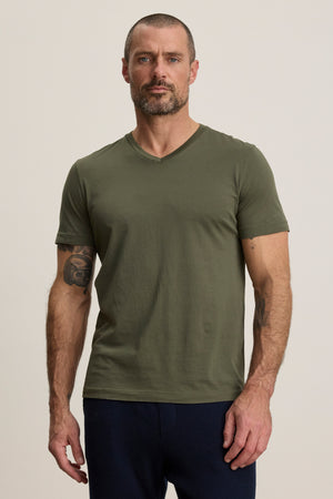 A man with short hair and a beard wears the olive green SAMSEN TEE by Velvet by Graham & Spencer and dark pants, standing against a plain light background.