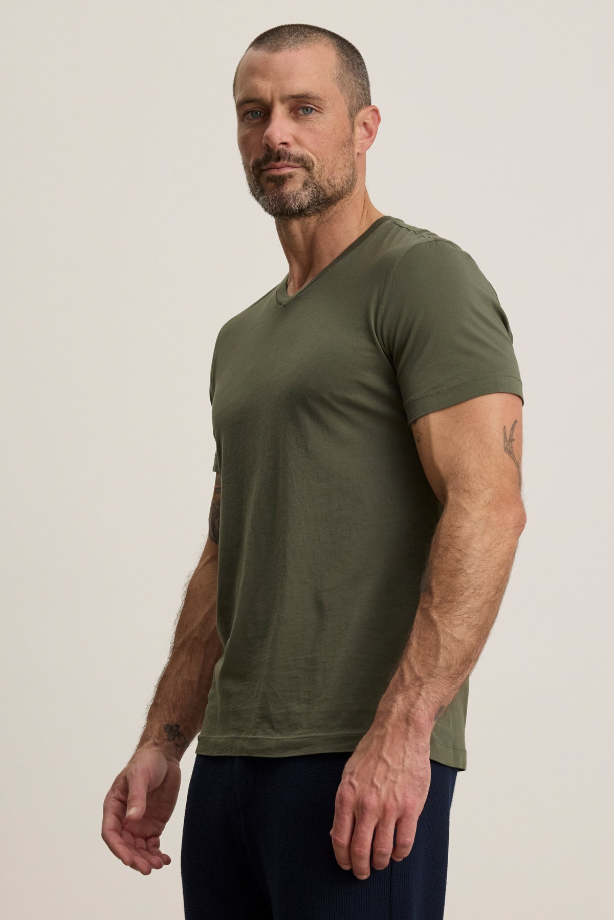A man with short hair and a beard models the SAMSEN TEE by Velvet by Graham & Spencer, an olive green v-neck tee paired with dark pants, standing against a plain light background—ideal for everyday wear.