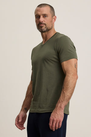 A man with short hair and a beard models the SAMSEN TEE by Velvet by Graham & Spencer, an olive green v-neck tee paired with dark pants, standing against a plain light background—ideal for everyday wear.