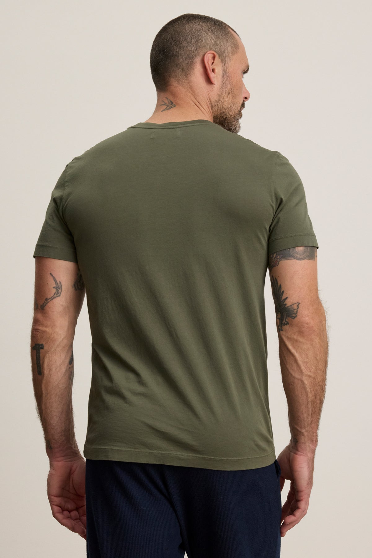 A man with short hair and tattooed arms stands facing away, wearing the Velvet by Graham & Spencer SAMSEN TEE in olive green—a Pima jersey v-neck tee paired with dark pants—an effortlessly stylish everyday look against a plain background.-40054351265985