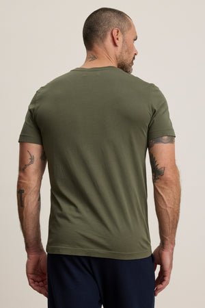 A man with short hair and tattooed arms stands facing away, wearing the Velvet by Graham & Spencer SAMSEN TEE in olive green—a Pima jersey v-neck tee paired with dark pants—an effortlessly stylish everyday look against a plain background.