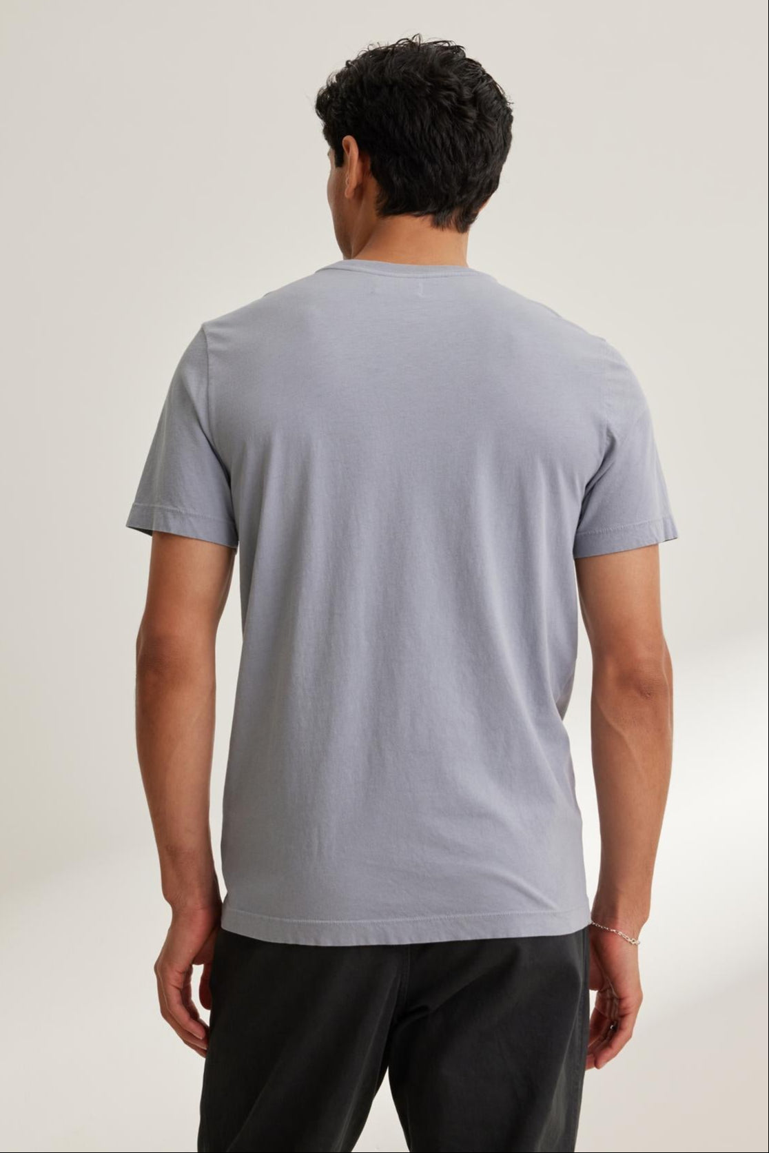 A person, with their back to the camera, wears a Velvet by Graham & Spencer SAMSEN TEE in light gray Pima jersey featuring a v-neckline, paired with dark pants against a plain background.